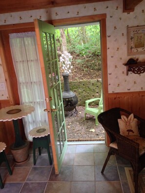 Une pièce chaleureuse avec une porte verte donnant sur un jardin, une petite table recouverte d’une nappe à motifs floraux, une chaise en osier et un poêle rustique.
