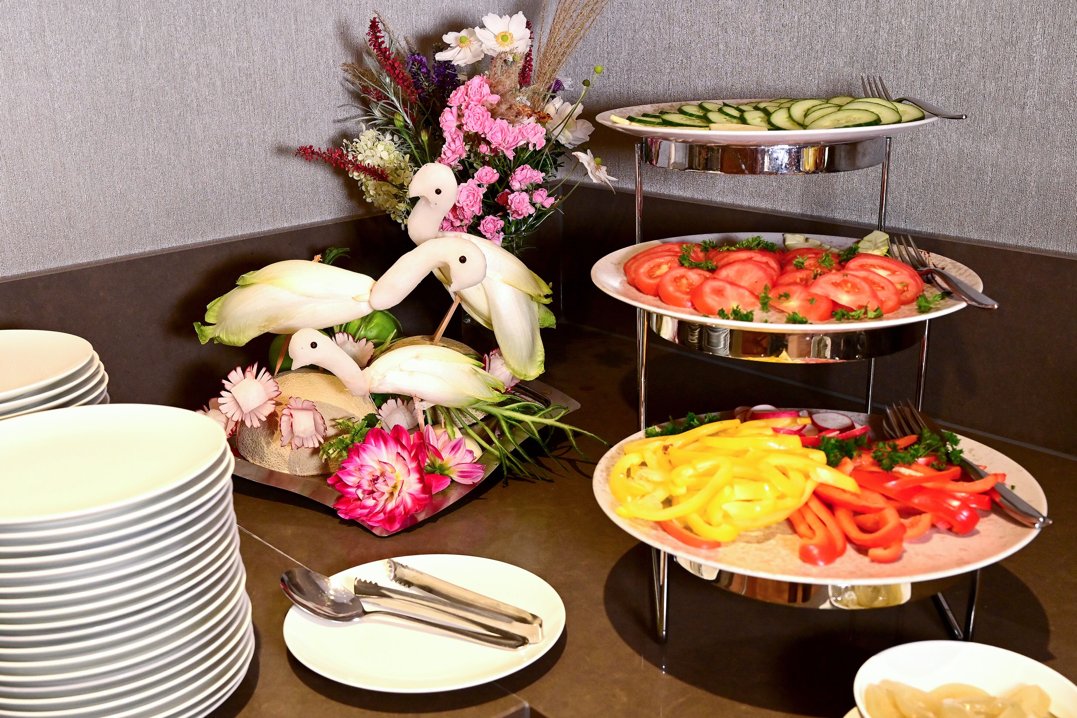 A buffet setup with various dishes, including sliced cucumbers, tomatoes, and bell peppers, arranged on a multi-tiered stand.