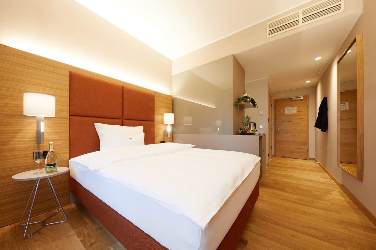 A hotel room with a large bed, two bedside lamps, a small table with a bottle and glass, a mirror, and a door leading to a hallway.