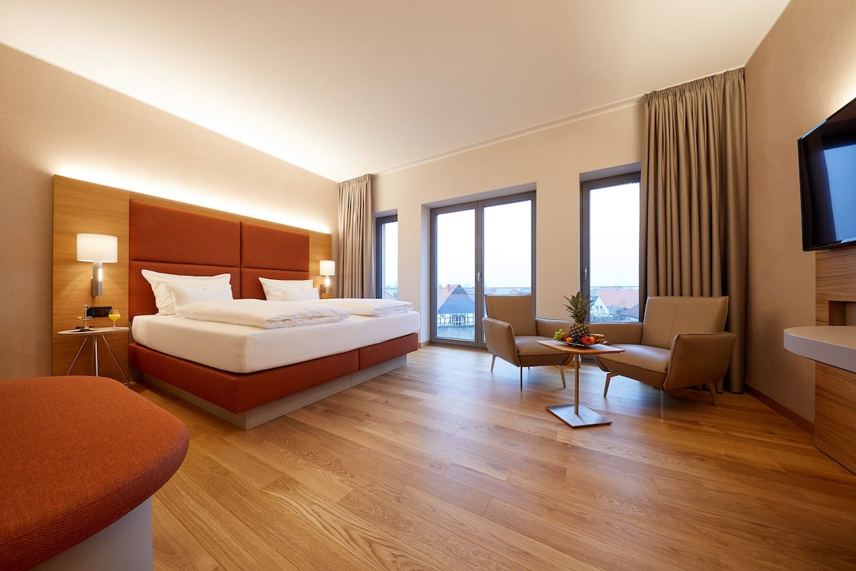 A hotel room with a large bed, a desk with a chair, a TV, and a balcony with curtains.