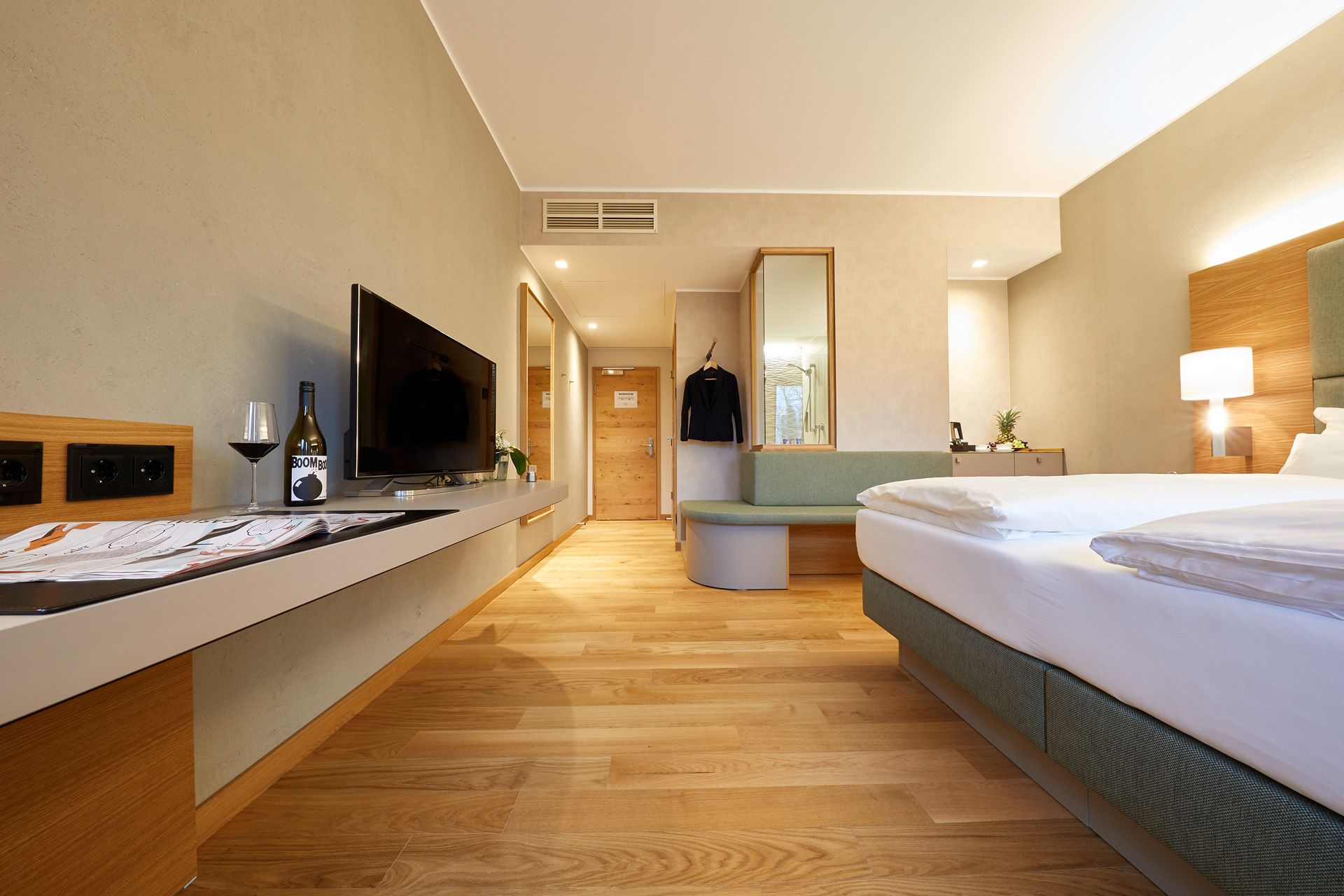 A modern hotel room with a large bed, a flat-screen TV, a minibar, a seating area, and a bathroom visible through an open door.