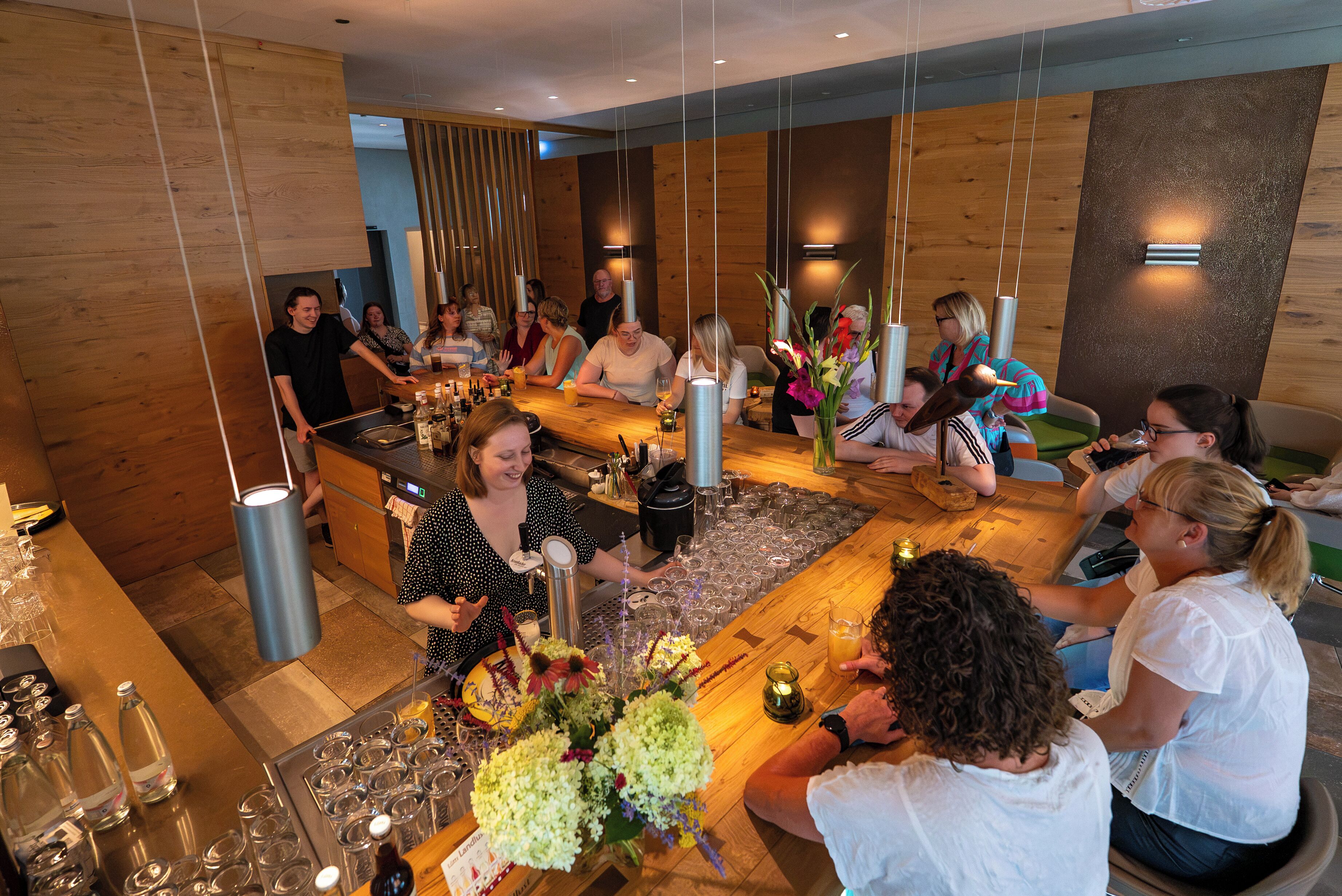 A group of people gathered around a bar area with a variety of bottles and glasses, indicating a social setting.