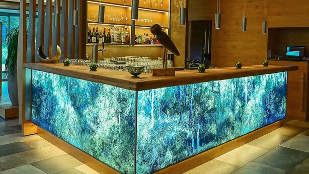 A bar area with a large illuminated screen displaying a forest scene, shelves with bottles, and glasses. - Hotel Rittergut Störmede (Geseke)