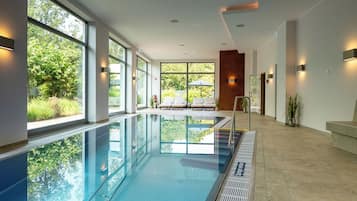 A modern indoor swimming pool with clear glass walls, a tiled floor, and a bench area with umbrellas.