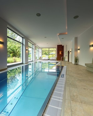 A modern indoor swimming pool with clear glass walls, a tiled floor, and a bench area with umbrellas.