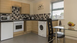 A compact kitchen with a microwave, oven, and washing machine, a glass table with a bowl of fruit, and a window with blinds. - Citystay Living - Dove House (Cambridge)