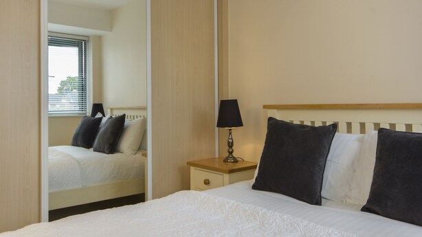 A hotel room with a bed, a nightstand with a lamp, a mirror, and a window with blinds. - Citystay Living - Dove House (Cambridge)