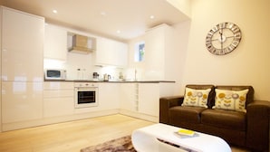 A modern kitchen with white cabinetry, a microwave, and a stove. There is a brown sofa with floral cushions and a white coffee table. A large wall clock is visible. - Citystay Living - Citygate (Cambridge)