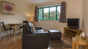 A modern living room with a leather sofa, a wooden coffee table, a TV on a wooden stand, a dining table with chairs, and a large window with curtains. - Citystay Living - Citygate (Cambridge)