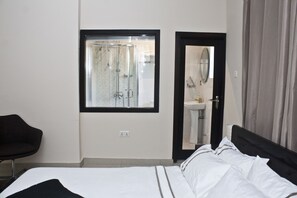A hotel room with a bed, a window with a view of a shower, a bathroom with a sink, and a chair. - Kegali Hotel (Accra)