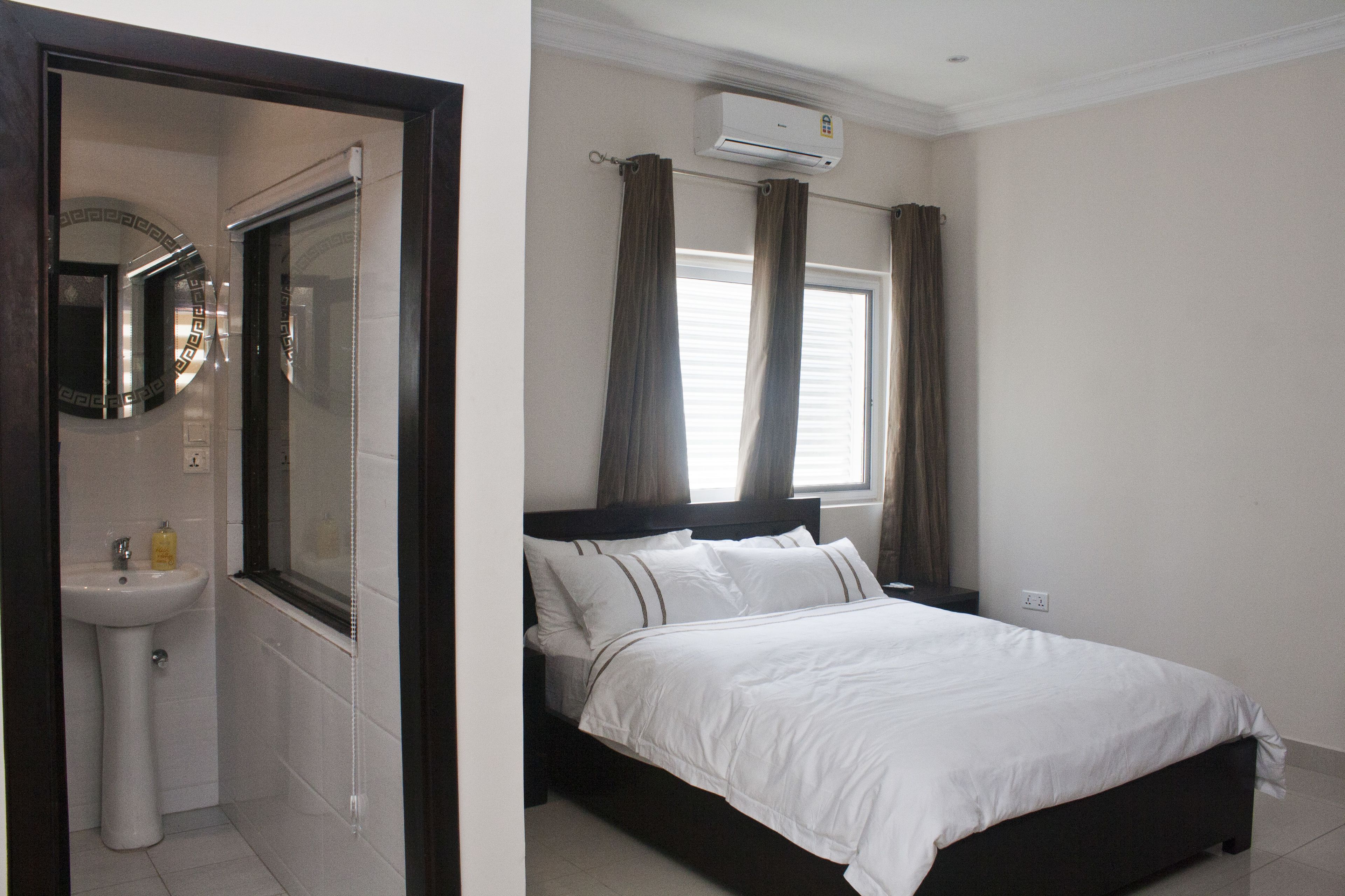 Een slaapkamer met een bed, een raam met gordijnen, een airconditioningapparaat en een badkamer met een wastafel en spiegel.