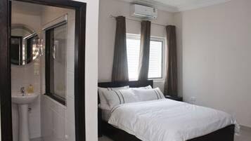 Een slaapkamer met een bed, een raam met gordijnen, een airconditioningapparaat en een badkamer met een wastafel en spiegel.