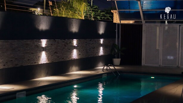 A swimming pool with a brick wall and a metal fence, illuminated by wall-mounted lights. - Kegali Hotel (Accra)