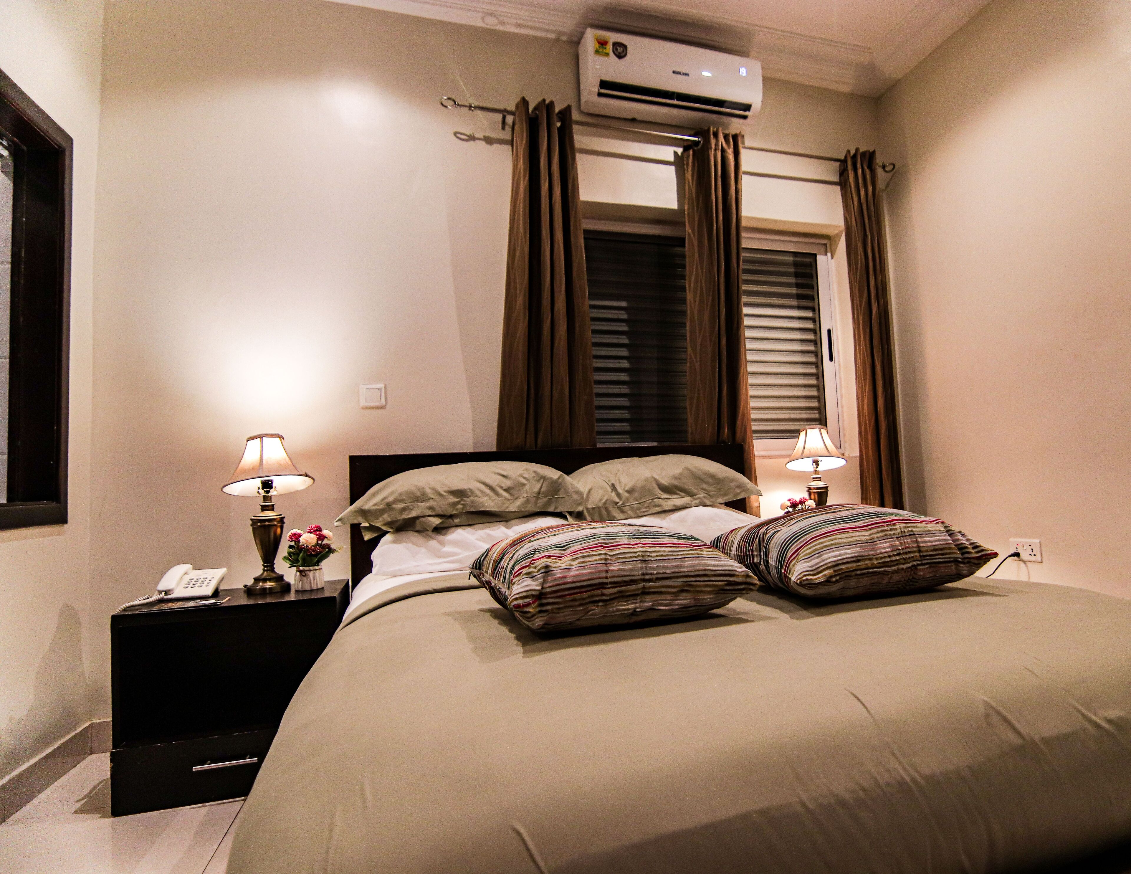 A bedroom with a bed, two bedside tables with lamps, an air conditioning unit, and a window with curtains.