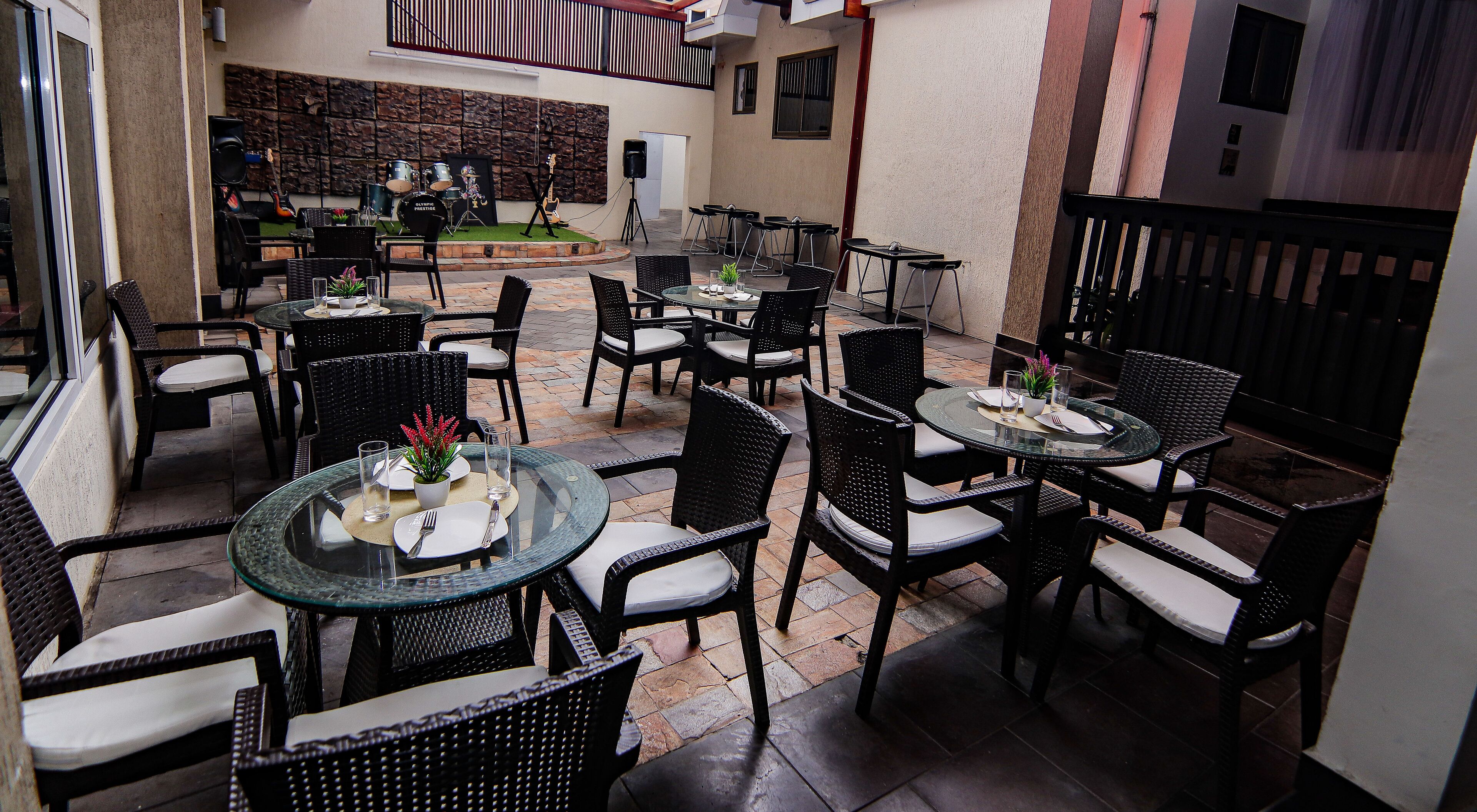 An outdoor patio with round tables and black wicker chairs, featuring a stone wall and a stage with musical instruments in the background.