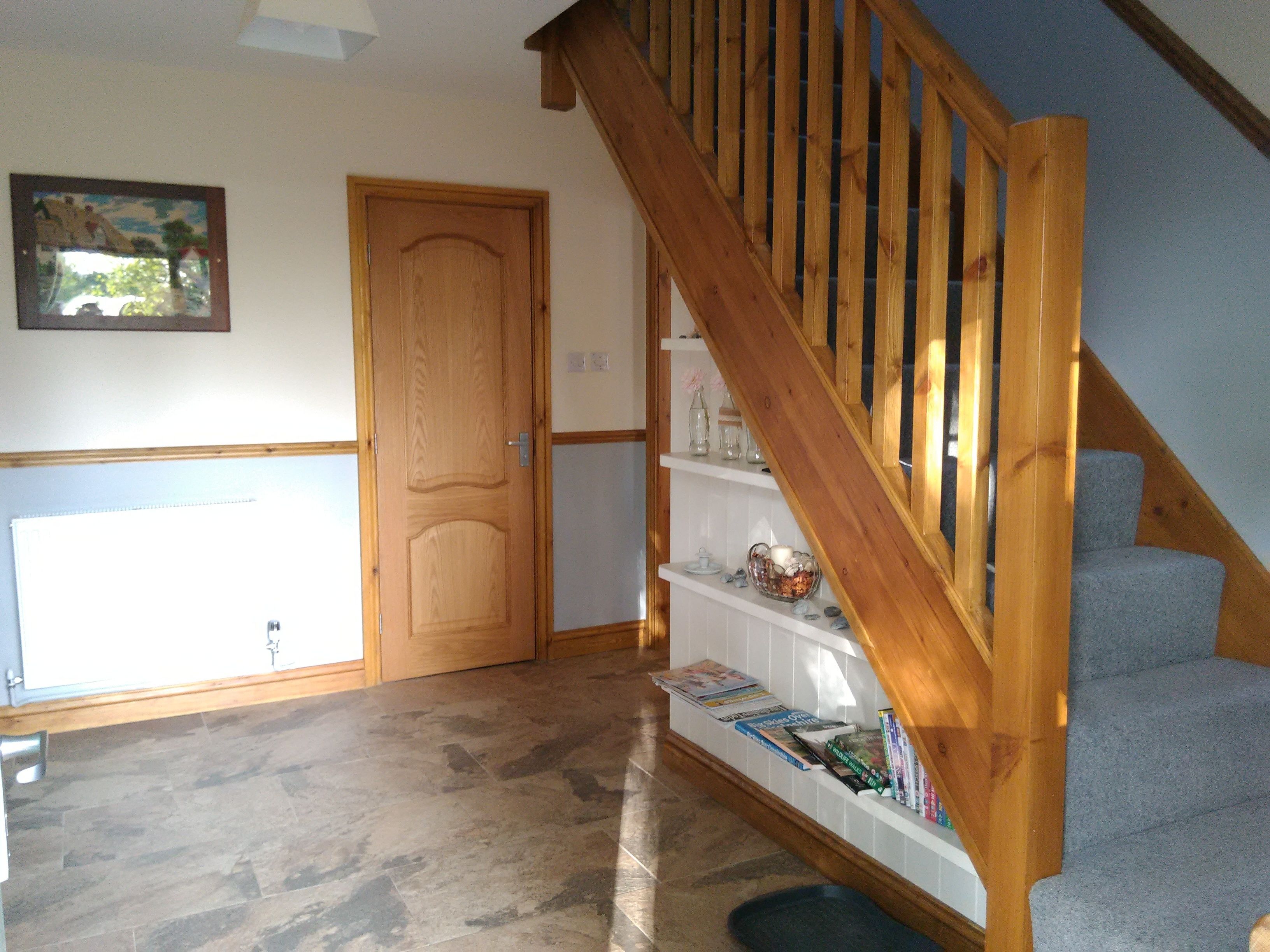 A hallway with a wooden staircase, a door, and a shelf with books and decorative items.