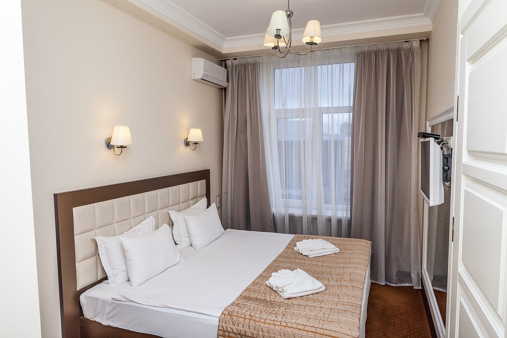 Habitación de hotel con una cama grande, dos lámparas de pared, un aire acondicionado, un televisor y una ventana con cortinas translúcidas.