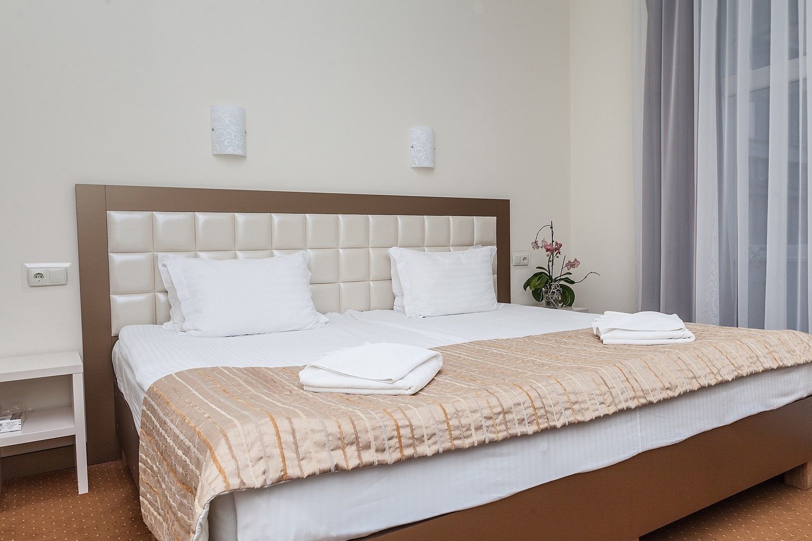 A neatly made bed with a beige headboard, white bedding, and a brown patterned throw.