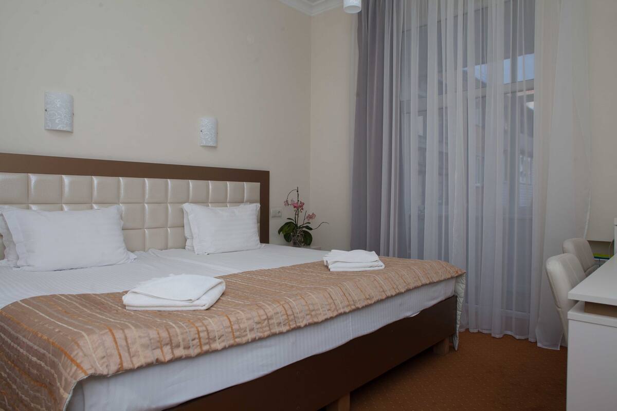 Una cama bien hecha con cabecera marrón, sábanas blancas y colcha estampada color beige. Hay una pequeña planta en maceta sobre la mesita de noche.
