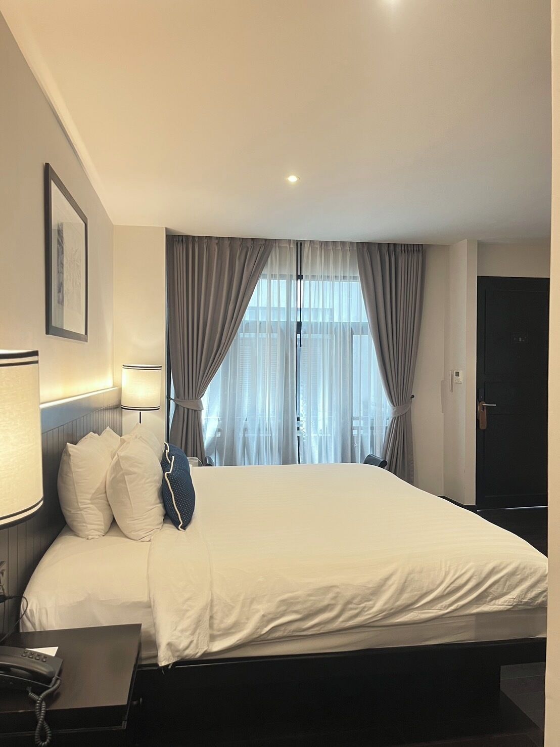 Una habitación de hotel con una cama grande, una mesita de noche con lámpara y una ventana con cortinas.