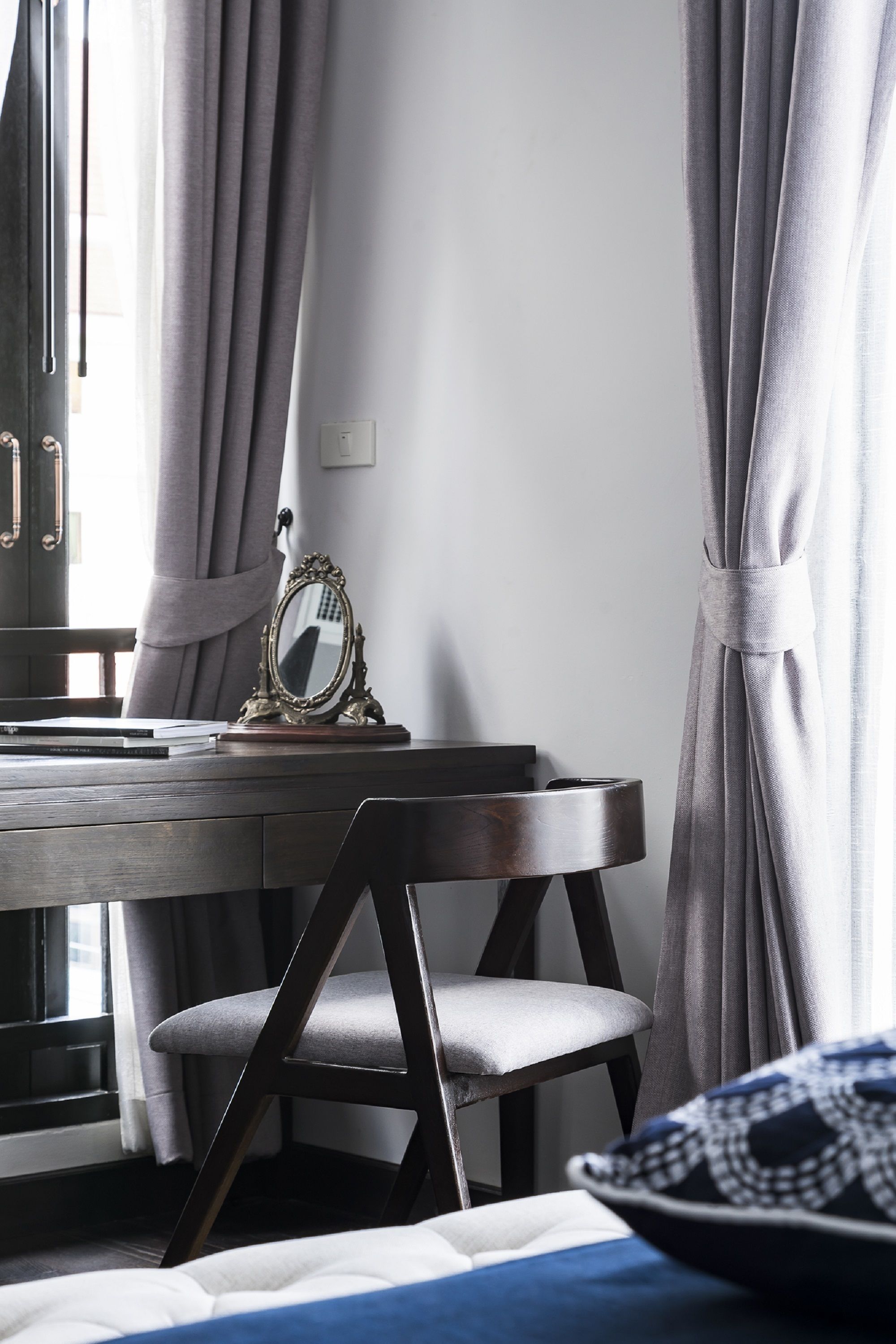 Ein Zimmer mit einem Holztisch, einem Stuhl, einem Spiegel und einem Bett mit blau-weißem Kopfkissen.