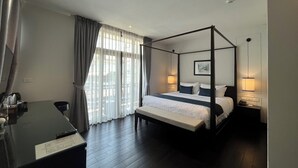 Una cama con dosel en una habitación de hotel, con escritorio y silla, ventanas amplias con cortinas y un cuadro en la pared.