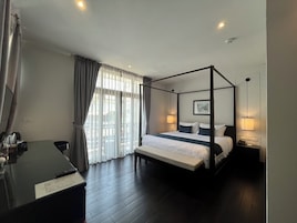 Una cama con dosel en una habitación de hotel, con escritorio y silla, ventanas amplias con cortinas y un cuadro en la pared.