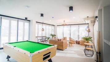 Una mesa de billar en una habitación luminosa con ventanas amplias, mobiliario de mimbre y un atril para billar.
