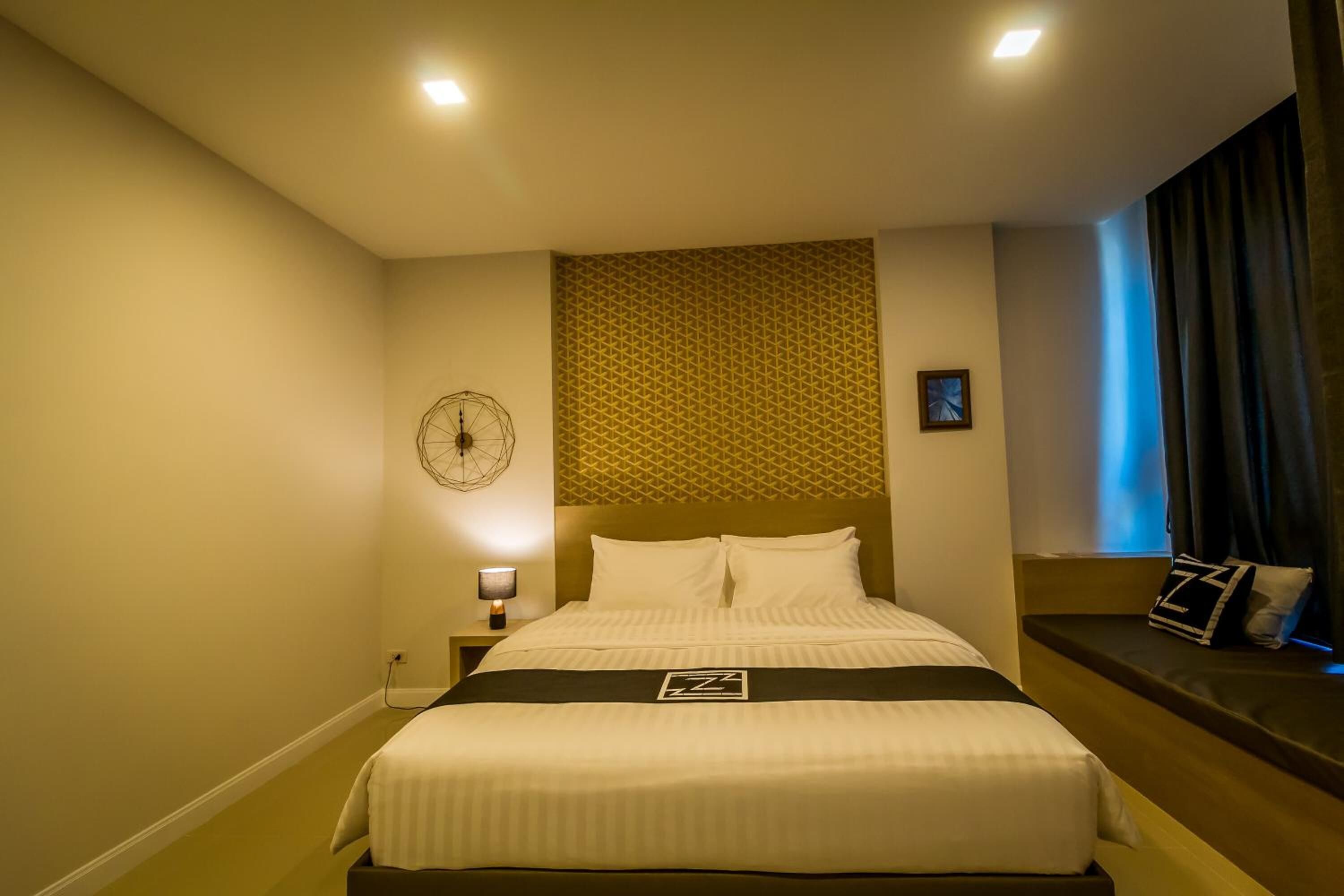 A hotel room with a large bed, a bedside table with a lamp, a wall clock, and a window with curtains.