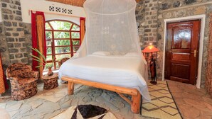 A room with a bed under a mosquito net, wicker furniture, a stone wall, a wooden door, and a window with red curtains. - The Palm Beach Resort (Nyayumba)