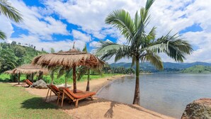 A beach with thatched-roof huts, palm trees, and lounge chairs. - The Palm Beach Resort (Nyayumba)