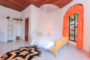 A room with a bed under a mosquito net, a wicker chair, a patterned rug, a white shelf with pottery, and a window with orange curtains. - The Palm Beach Resort (Nyayumba)