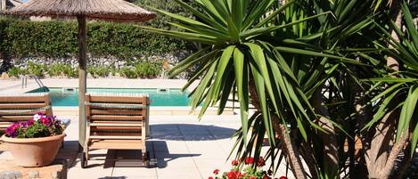 A poolside area with a thatched umbrella, wooden lounge chairs, and a potted plant with flowers.