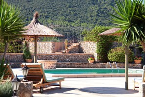 A pool area with lounge chairs, a thatched-roof gazebo, and a stone wall. - Ca's Garriguer Agroturismo (Valldemossa)