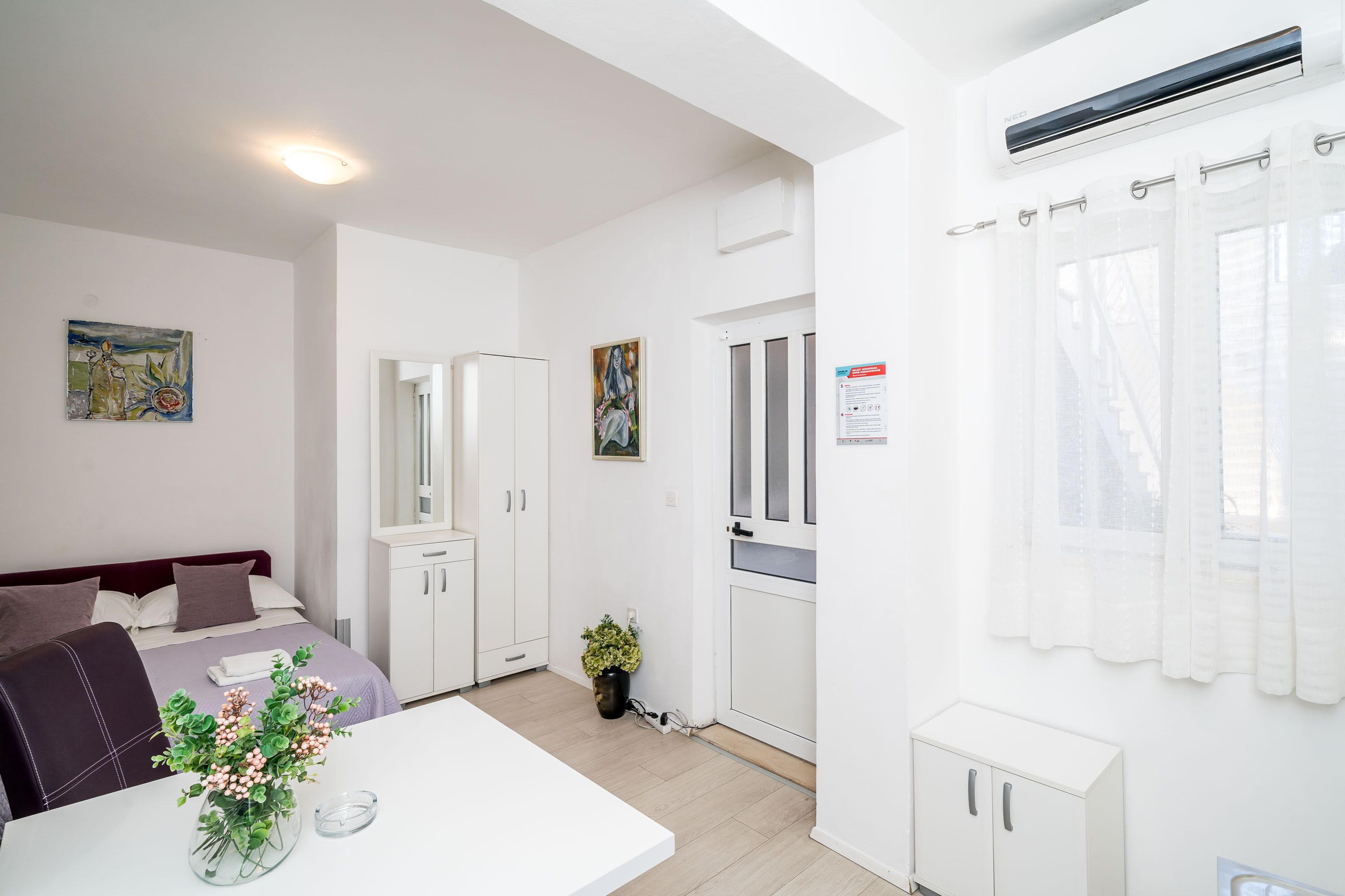 A modern hotel room with a bed, a dining table with a vase of flowers, a kitchen area, and a bathroom.