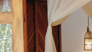 A room with a bed, a wooden nightstand, a vase, a hat, and a window with a view of greenery.