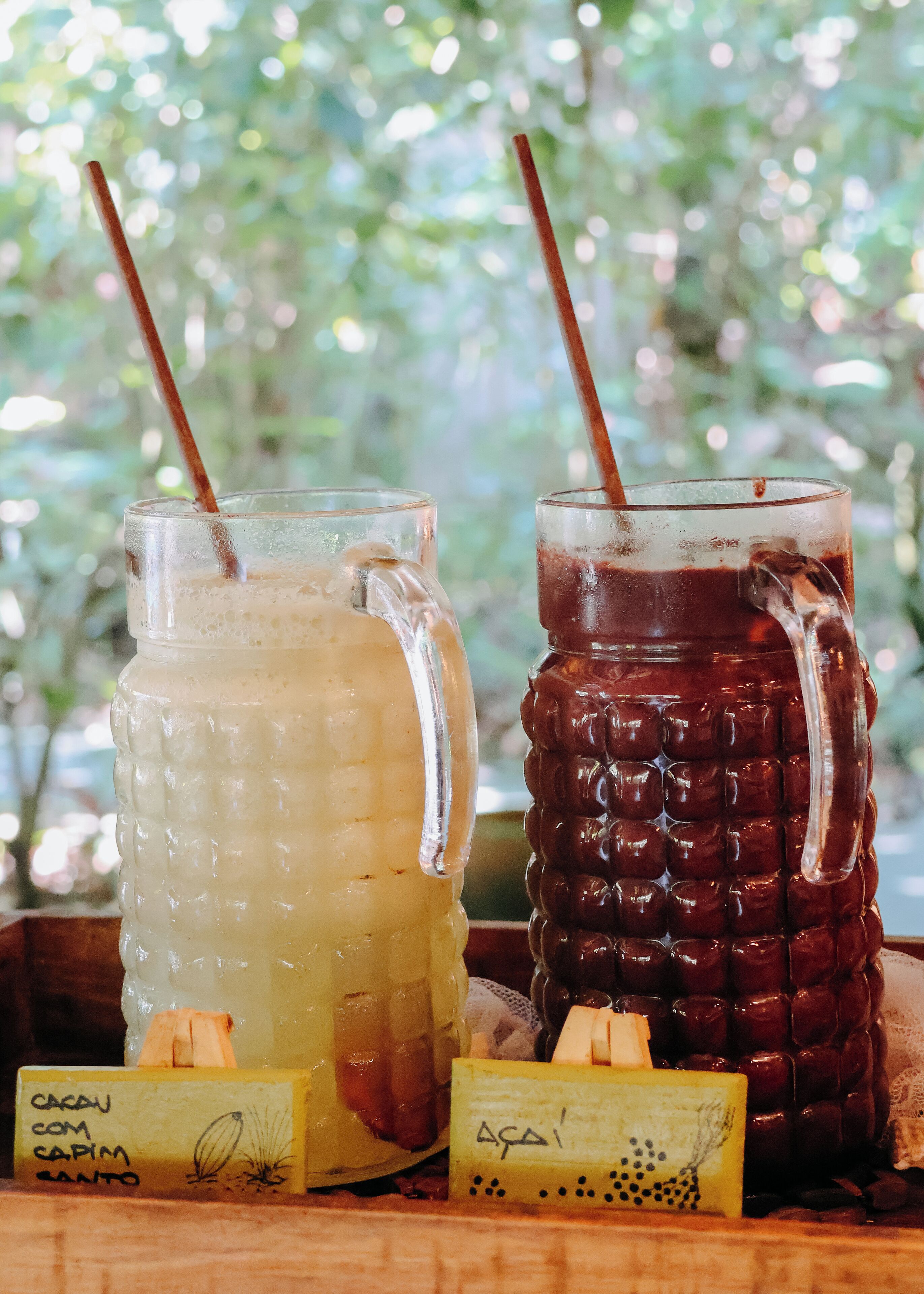 Two glass containers with straws, one filled with a light-colored beverage and the other with a dark beverage, placed on a wooden surface with labels.