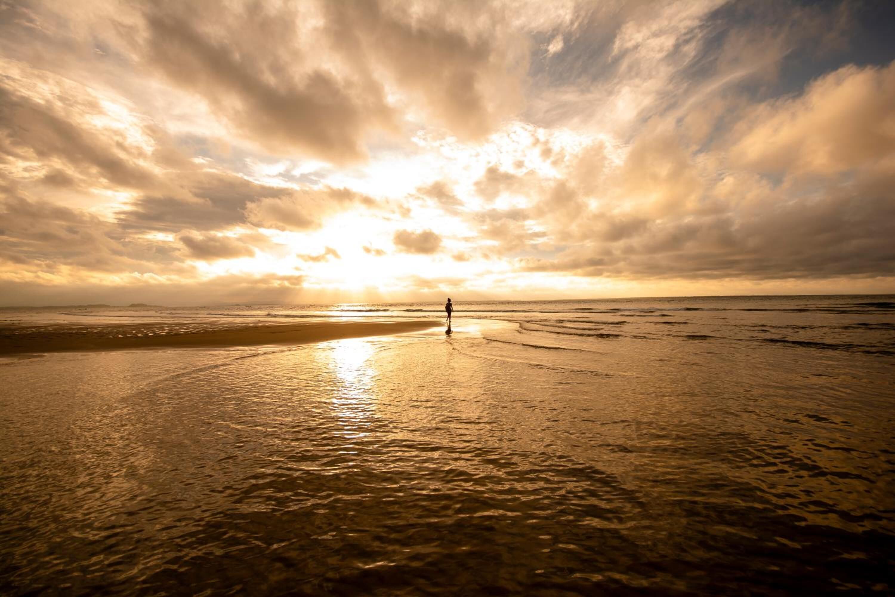 A beach with a person standing in the middle, facing the sunset.