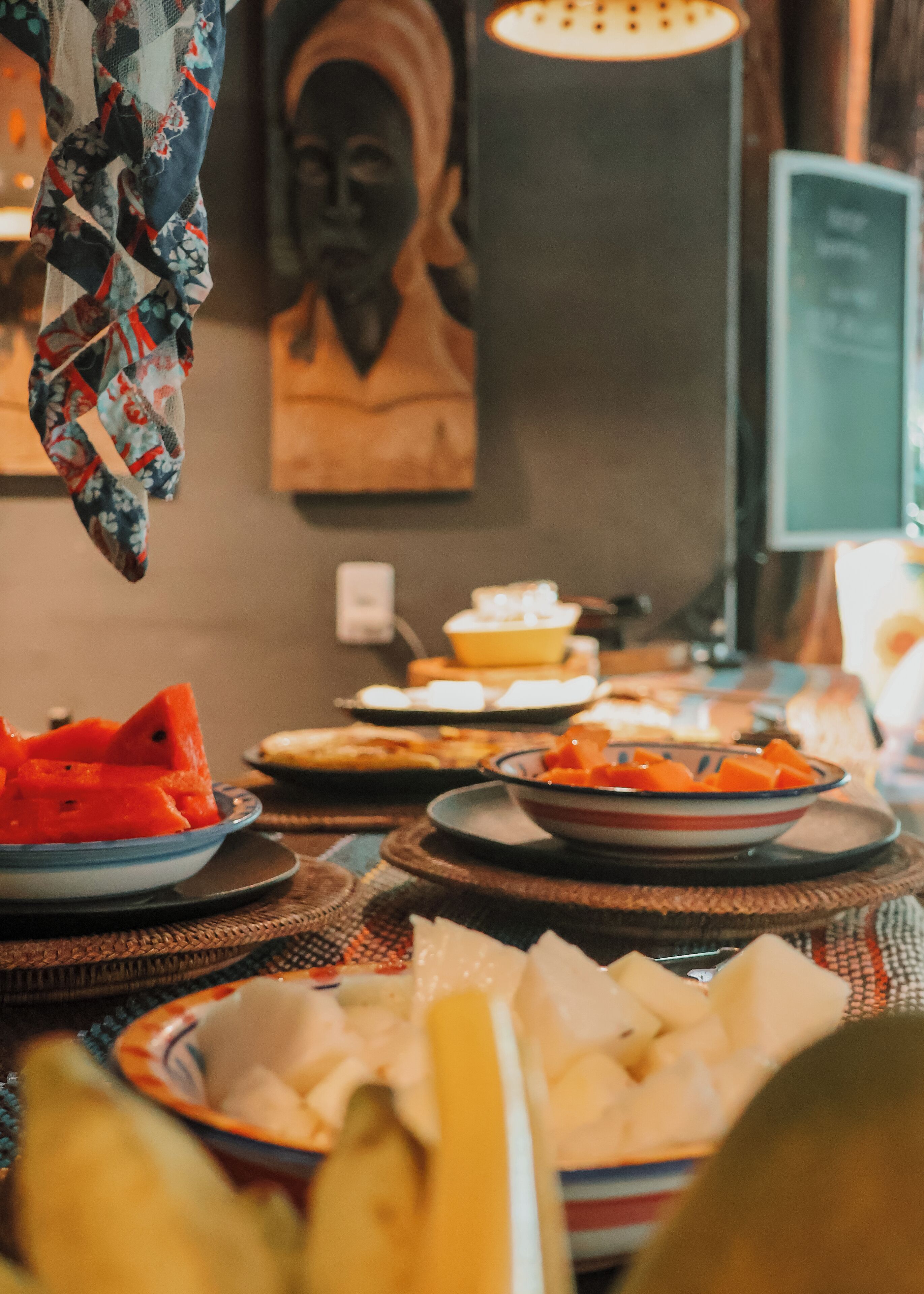A dining table with various dishes, including watermelon and cheese, in a room with a wall-mounted head sculpture and a computer monitor.