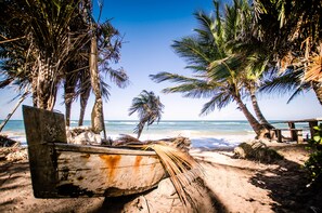 A beach with palm trees, a wooden boat, and a wooden walkway. - Na Villa dos Algodões (Maraú)