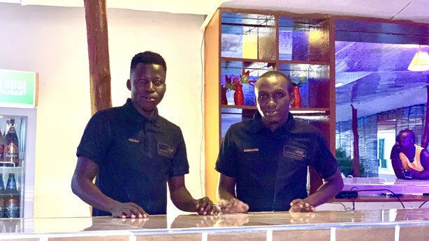 Two individuals standing behind a counter with a brick facade, a sign indicating a restaurant, and a reflection of a person in the background. - Sundowner Lodge (Tamale)