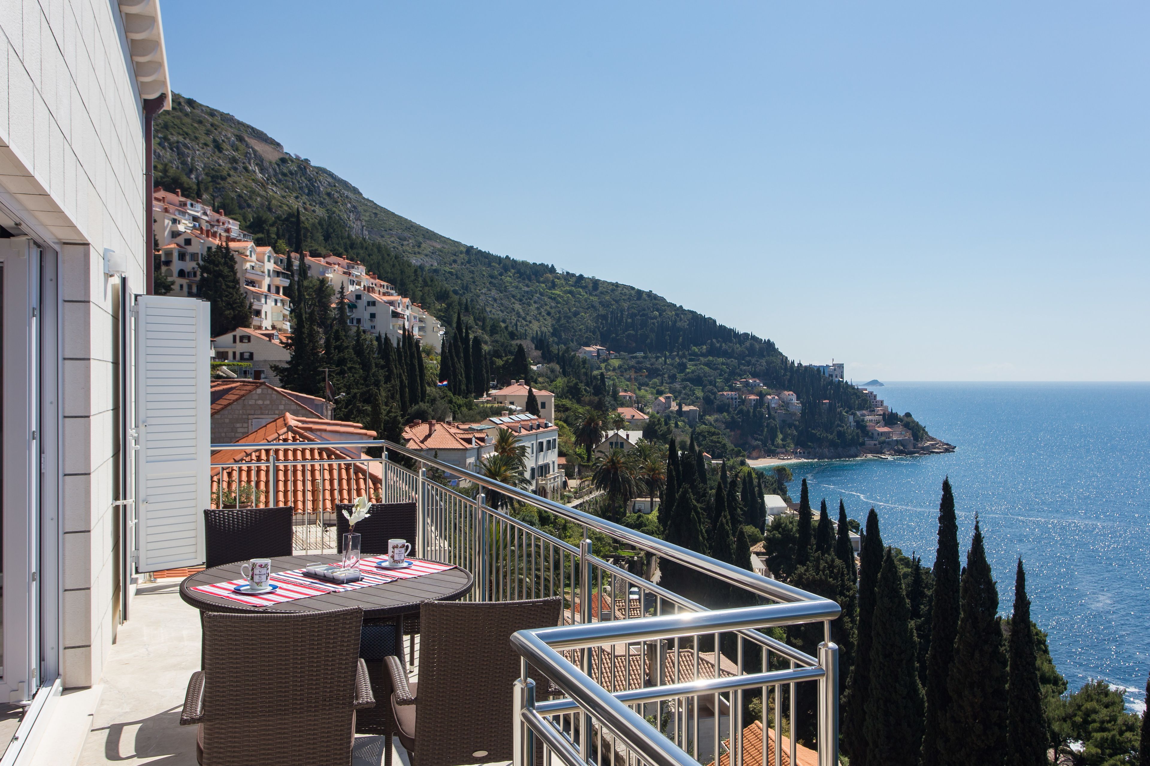 A balcony with wicker furniture and a table, overlooking a coastal town with buildings and a clear blue sky.