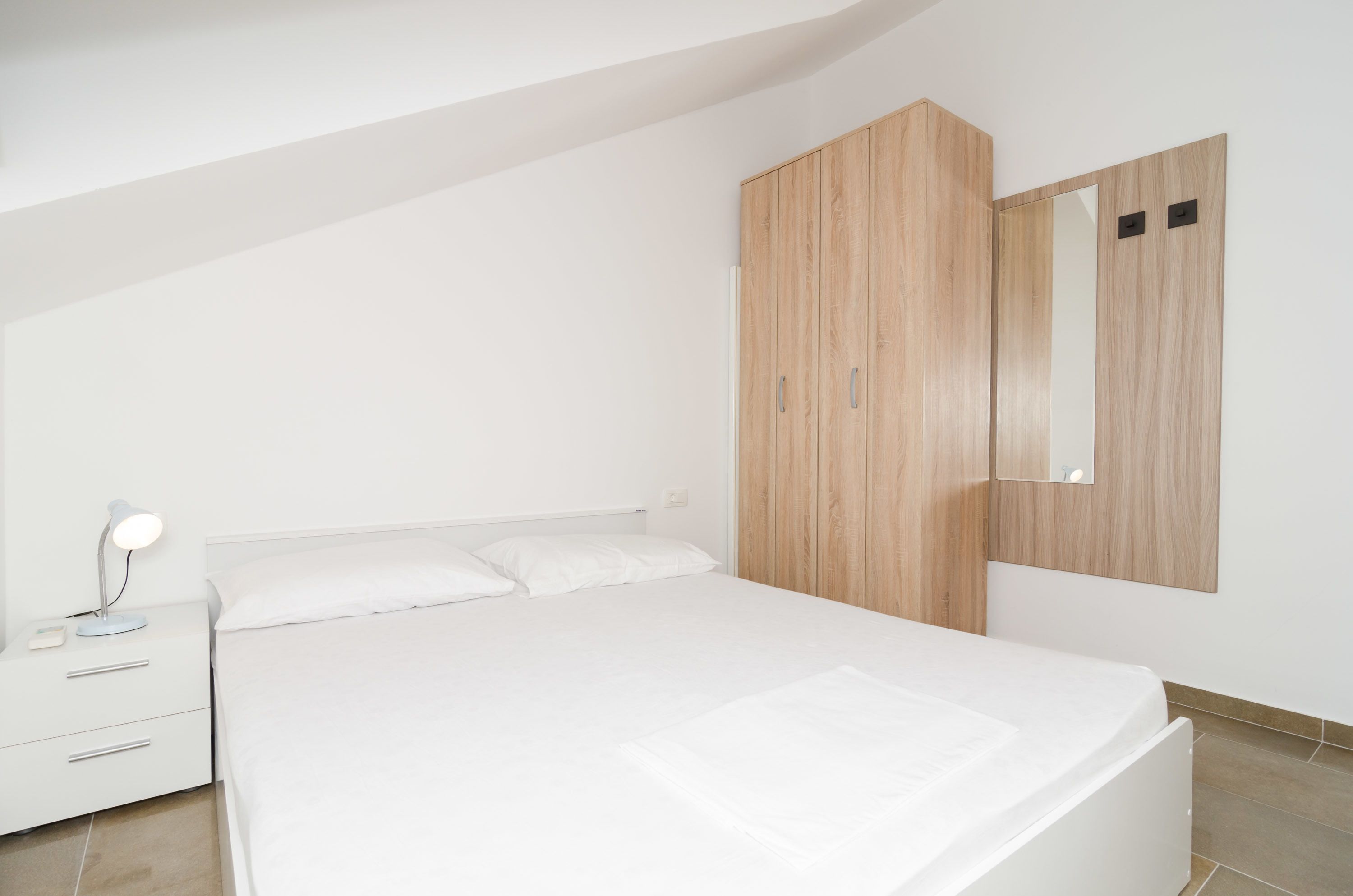 Een moderne slaapkamer met een bed, een nachtkastje, een bedlamp en een houten garderobe met spiegel.