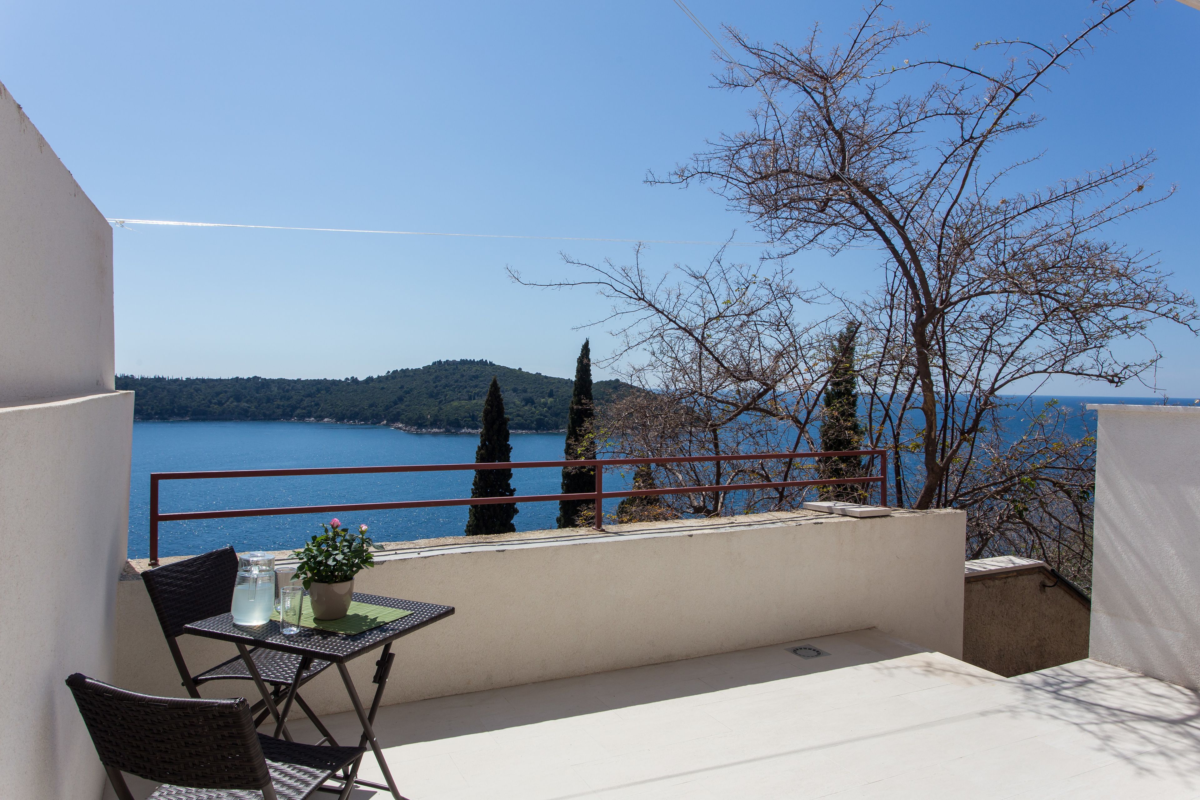 A balcony with a table, chairs, and a potted plant overlooking a body of water and hills.
