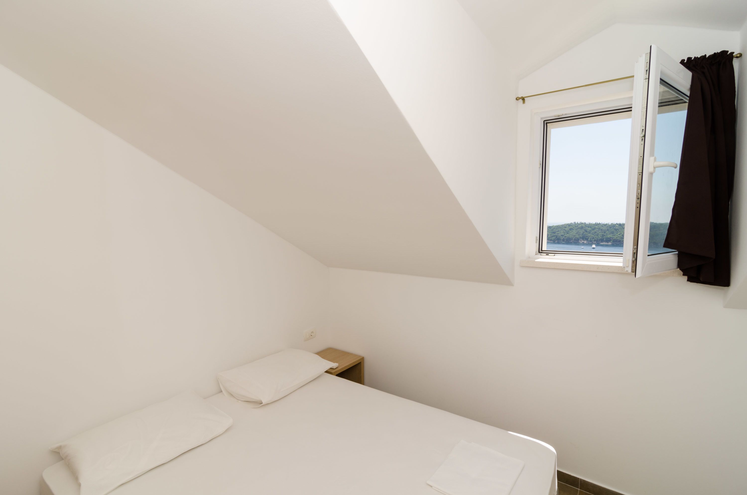 A small, white, single-bed room with a sloped ceiling and a window with a curtain.