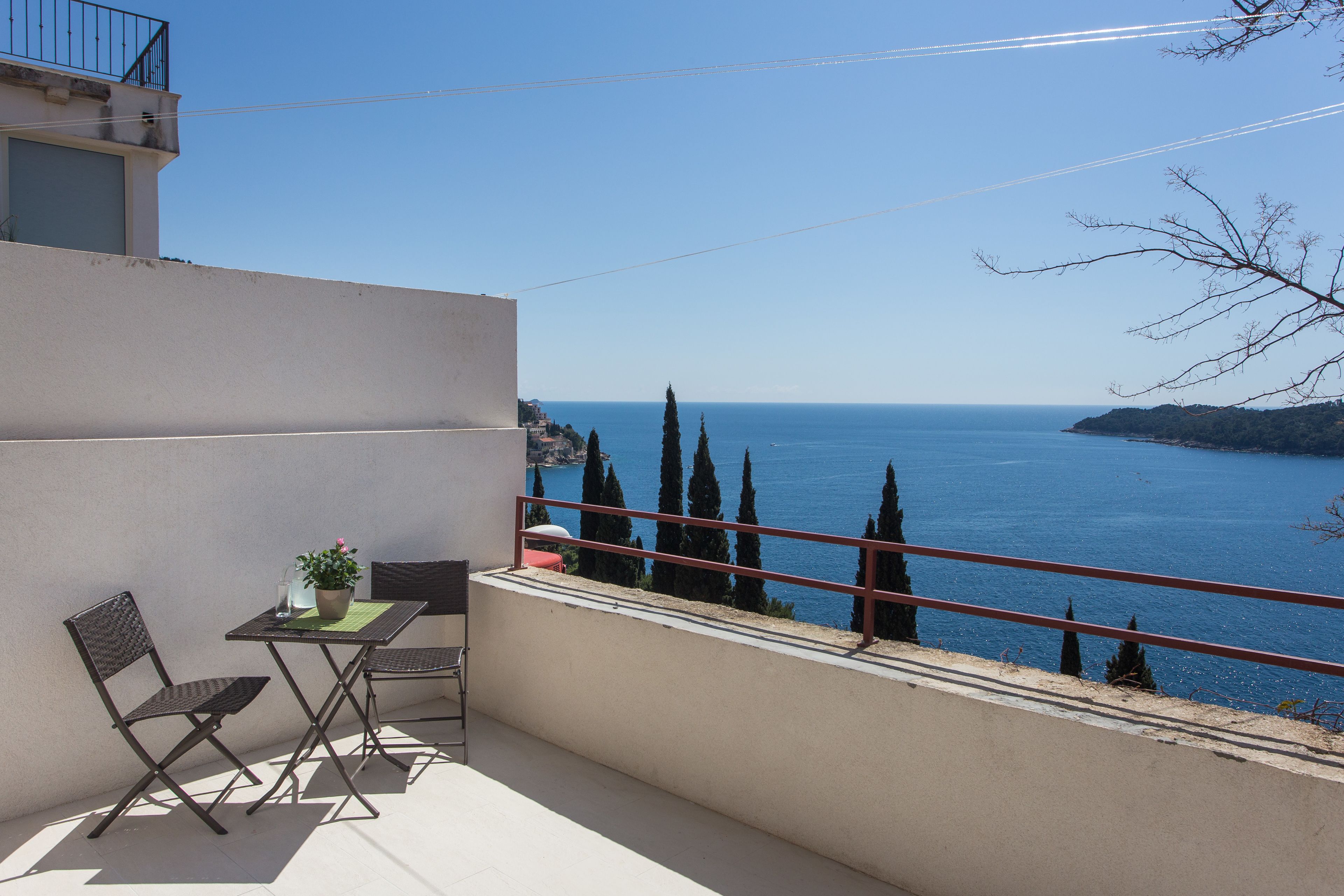 Un balcón con mesa y sillas, con vistas al mar y cipreses.