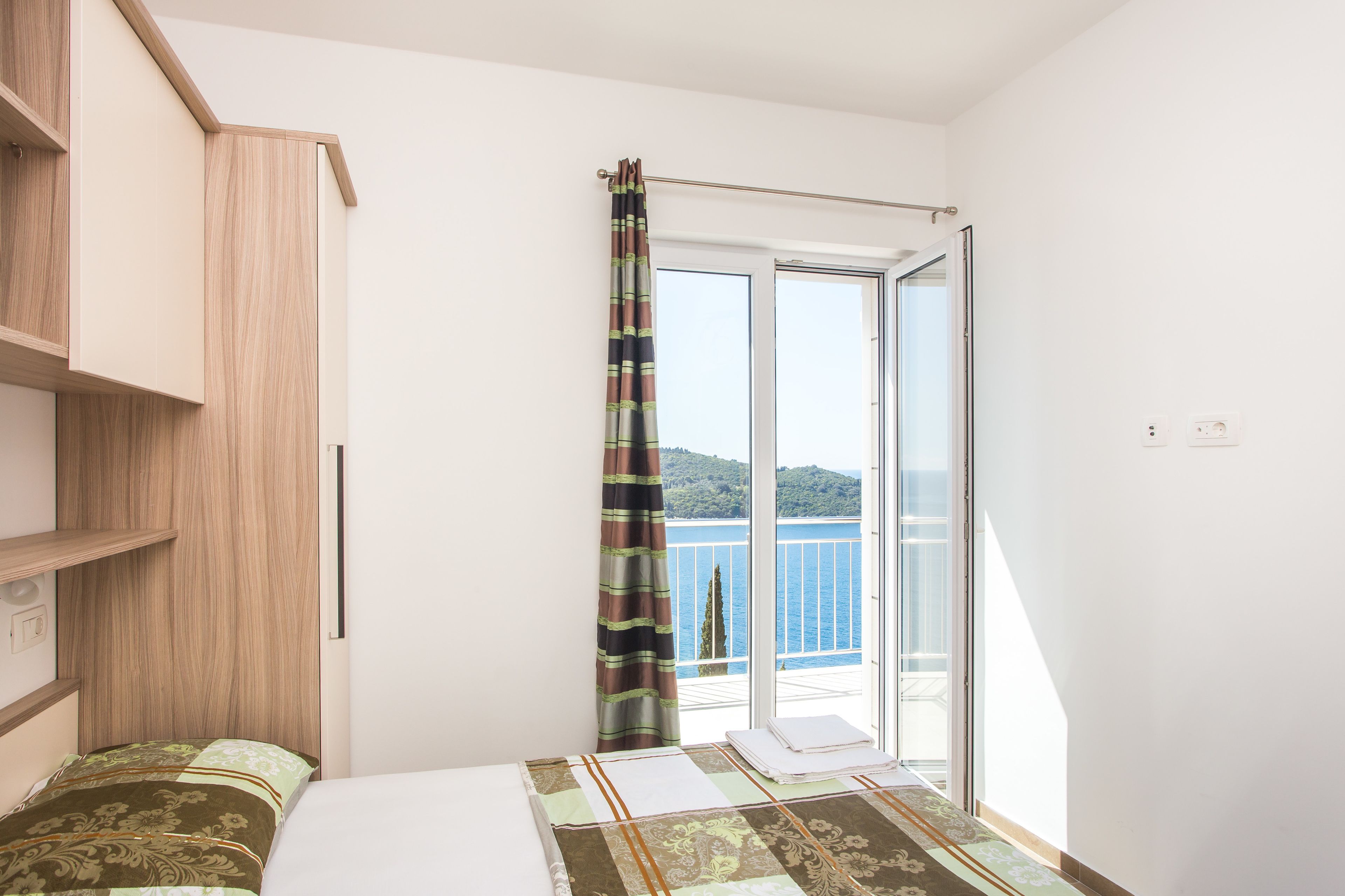 A bedroom with a bed, a wooden wardrobe, a window with a view of the sea, and a balcony with a curtain.