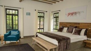 A bedroom with a large bed, wooden headboard, and a bench. There are two windows with curtains, a chair, and a small table. - Arusha Villa (Arusha)