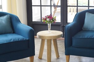 Dos sillones azules con cojines, una mesita de madera con un jarrón de flores, y vistas a través de una ventana.