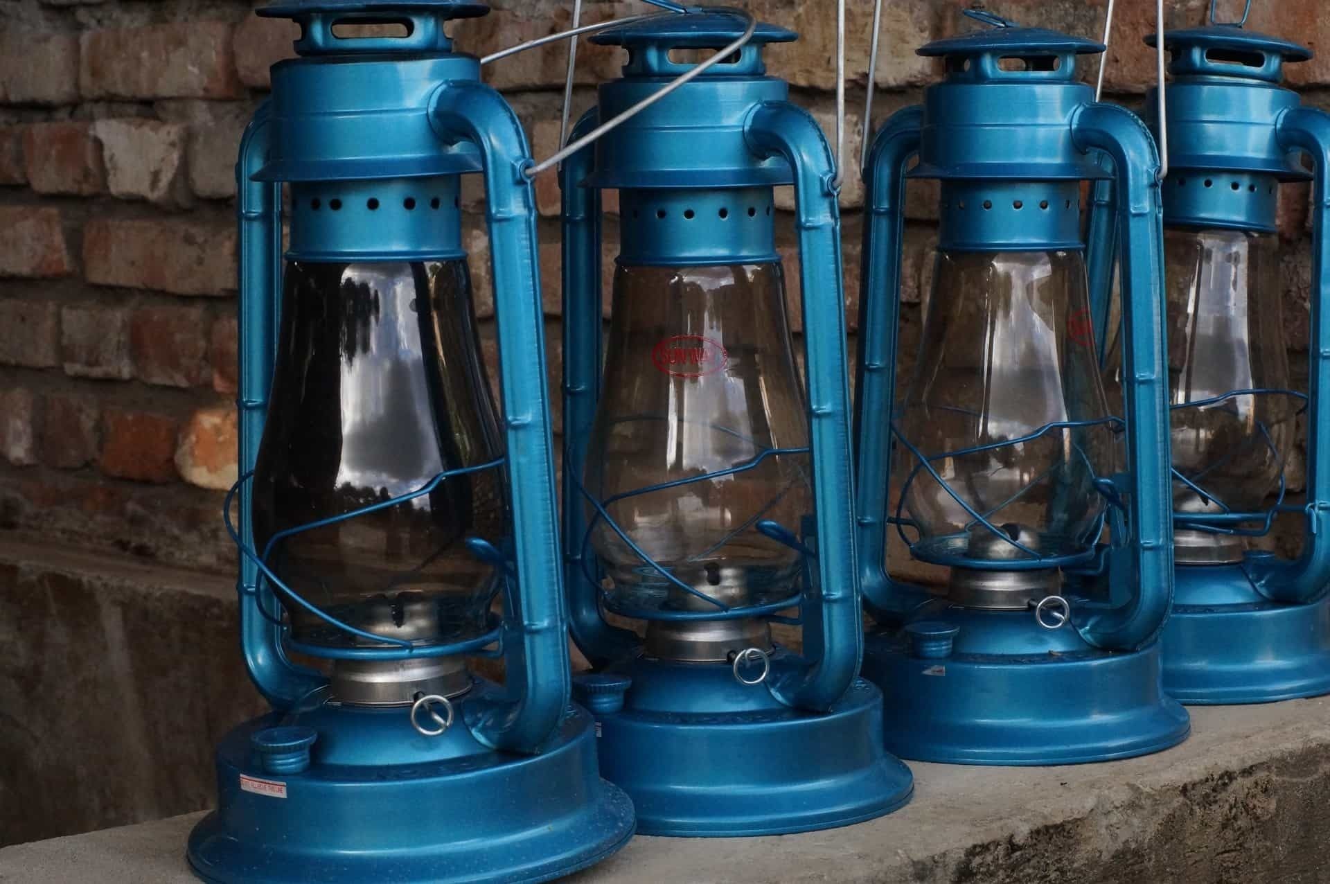Vier blauwe lantaarns met glazen omhulsels en metalen voetstukken, in een rij voor een bakstenen muur.